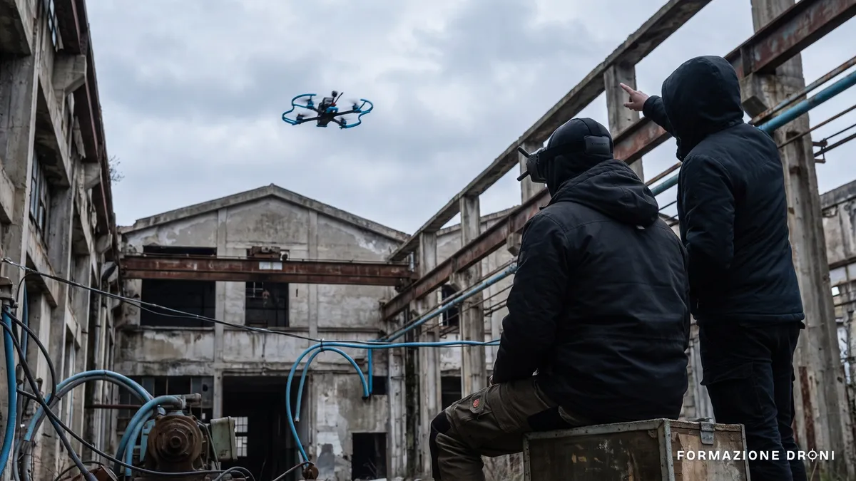 Pilota seduto con visore FPV e osservatore accanto che segue il drone in volo in un'area industriale dismessa