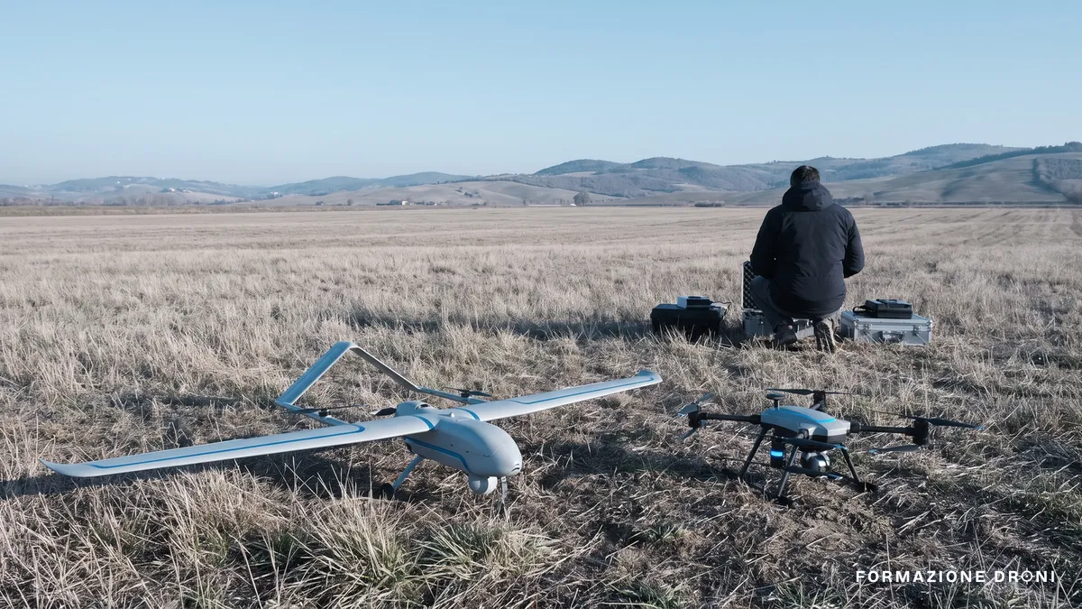 Un drone ad ala fissa e un quadricottero appoggiati fianco a fianco su un prato, con pianura agricola italiana sullo sfondo